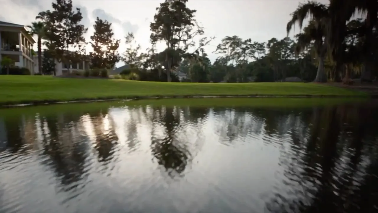 Late afternoon light across a lawn and lagoon, live oaks and houses beyond. A coastal Georgia house at sunset, its windows lit above the water and marsh.